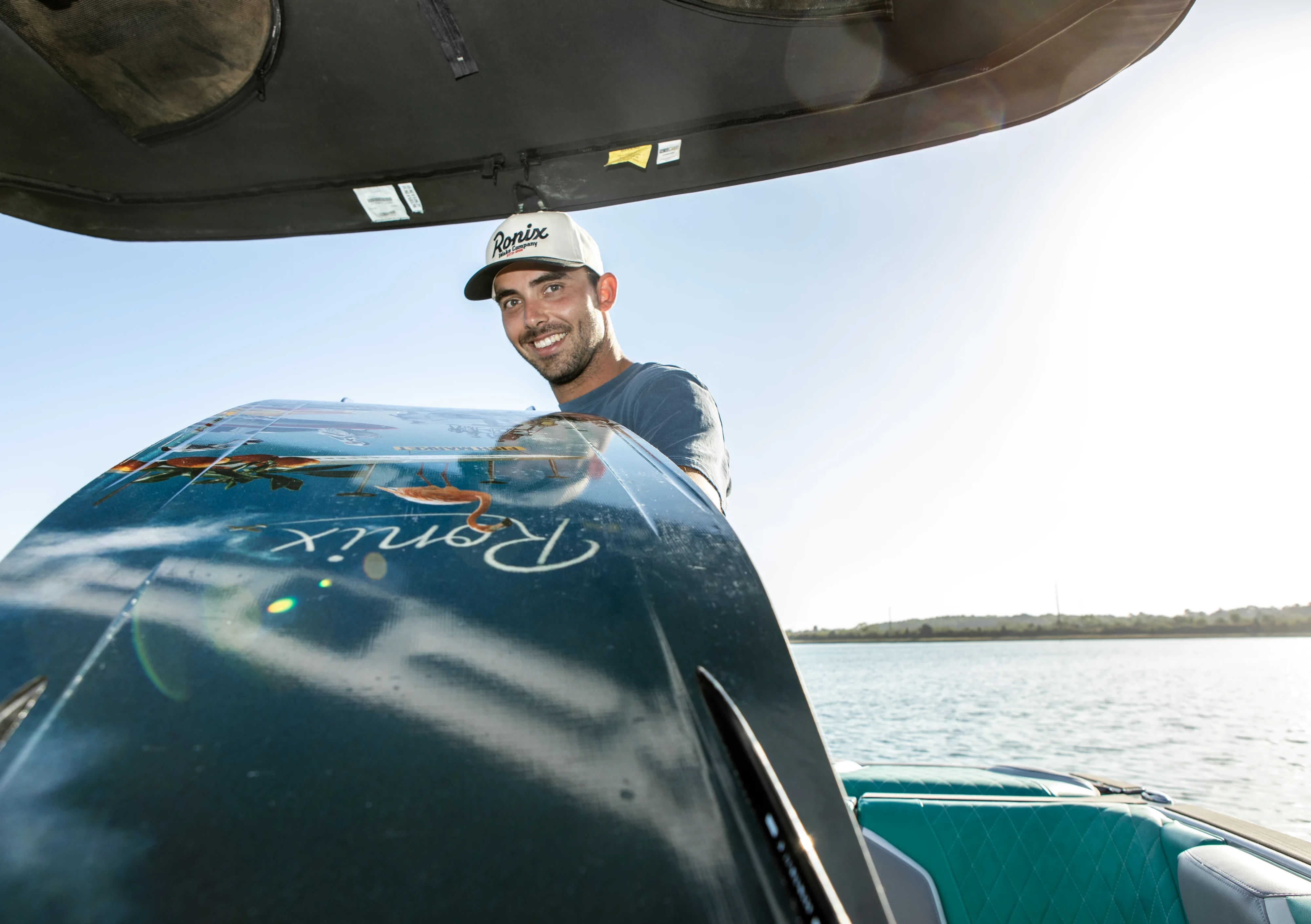 Lifestyle shot of Jake Pelot holding the Ronix Supreme wakeboard, showing off the base graphic, with water and boat in the background